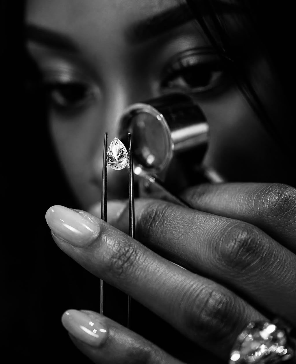 A close-up black and white photograph of a person holding a small clear gemstone between tweezers, with a jeweler's loupe near the eye, and a ring on one finger.