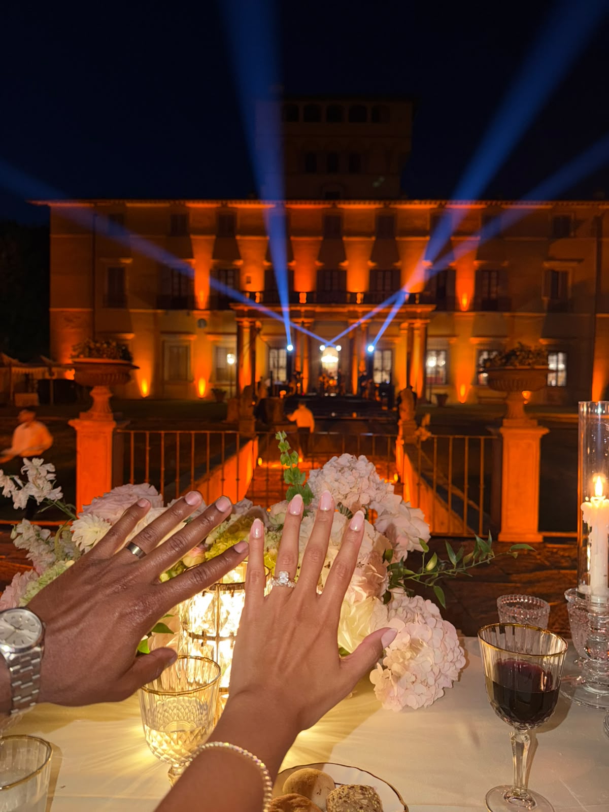 A couple's hands raised in celebration at a dinner table, wearing Novia Diamonds bespoke matching rings, with a beautifully lit villa in the background at night under blue spotlight beams.